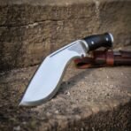 1095 steel kukri with black micarta grip and knotwork bolster, low angle close view of the upright blade on a sunlit stone step