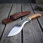1095 steel kukri with olive wood grip and Mjolnir cut brass bolster, closer angled view of the blade on weathered deck boards