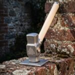 Hand forged blacksmith hammer with an octagonal 1095 carbon steel head, engraved Valknut on the cheek and an American ash handle, standing on a weathered brick wall