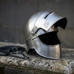 Brushed steel visored medieval helmet with valknut brow engraving and perforated bevor resting on a stone bench beside its black leather liner