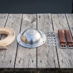 Hand raised steel Viking shield boss with engraved Valknut laid on weathered wood beside leather rim binding, rivets, washers and brown leather arm straps