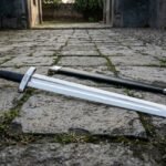 Hand forged norse warrior sword with a broad fullered polished blade, black leather grip and lobed steel pommel lying on a cobbled street beside its black scabbard