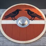 Viking shield of planked wood painted with twin black ravens on a red and white field, steel valknut boss centred, leather strap and grips alongside.