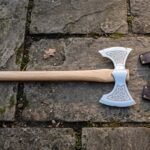 Hand forged double bit axe with etched Celtic knotwork on both flared bits and a pale ash haft, laid on mossy flagstones beside a brown leather head cover.