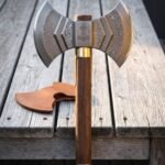 Hand forged Damascus double headed axe with layered twin crescent bits, an engraved Mjolnir cheek, walnut haft and brass collar standing on a weathered wooden deck