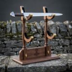 Walnut sword display stand with brass lined cradles and a cut Mjolnir engraving holding a Viking sword, photographed on a stone ledge before a dry stone wall