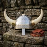 Hand forged horned Viking helmet with a riveted steel dome, brass rivets and trim, engraved knotwork browband, nasal bar and pale horns, on a dry stone wall.