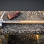 Viking war hammer with a forged square face, curved beak and Mjolnir engraved head on a pale ash haft with steel langets, lying on stone beside its leather head cover