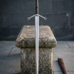 Hand forged damascus longsword with a leather wrapped grip and quatrefoil cross guard standing point down beside a cross laced leather scabbard on grey stone