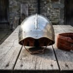 Hand forged Viking helmet with a riveted steel dome, brass rivets, an engraved brass browband and a brown suede liner, on weathered timber outside a stone building.