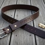 Hand made dark brown full grain leather rune belt stamped with an Elder Futhark line, brushed steel buckle and brass rivets, coiled on weathered grey decking