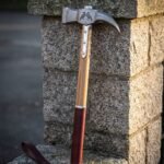Hand forged war hammer with a Damascus steel head, etched ravens and a curved fluke, ash haft with steel langets and an oxblood leather grip, against a granite pillar.