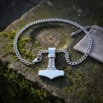 Polished stainless steel mjolnir pendant with a Celtic knotwork bail and scrolled shaft on a heavy curb chain, resting on mossy stone beside a grey gift pouch