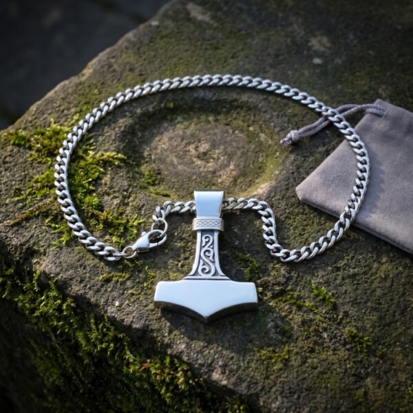 Polished stainless steel mjolnir pendant with a Celtic knotwork bail and scrolled shaft on a heavy curb chain, resting on mossy stone beside a grey gift pouch