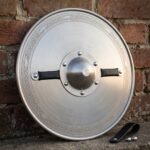 Hand forged steel round shield with a dished polished face, etched knotwork border, riveted dome boss and black leather grip, resting against a brick wall.