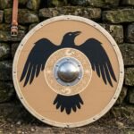 Hand made viking round shield with a painted black raven, engraved knotwork roundel, steel dome boss and rawhide rim, leaning on a dry stone wall