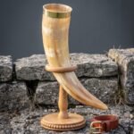 Valknut drinking horn in pale honey ox horn with a solid brass rim band, held in a turned oak ring stand beside a tan leather belt