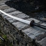 Hand forged katana sword with a mirror polished 1095 blade, bo hi groove, round iron tsuba and black cord wrapped handle on a dry stone wall beside its saya