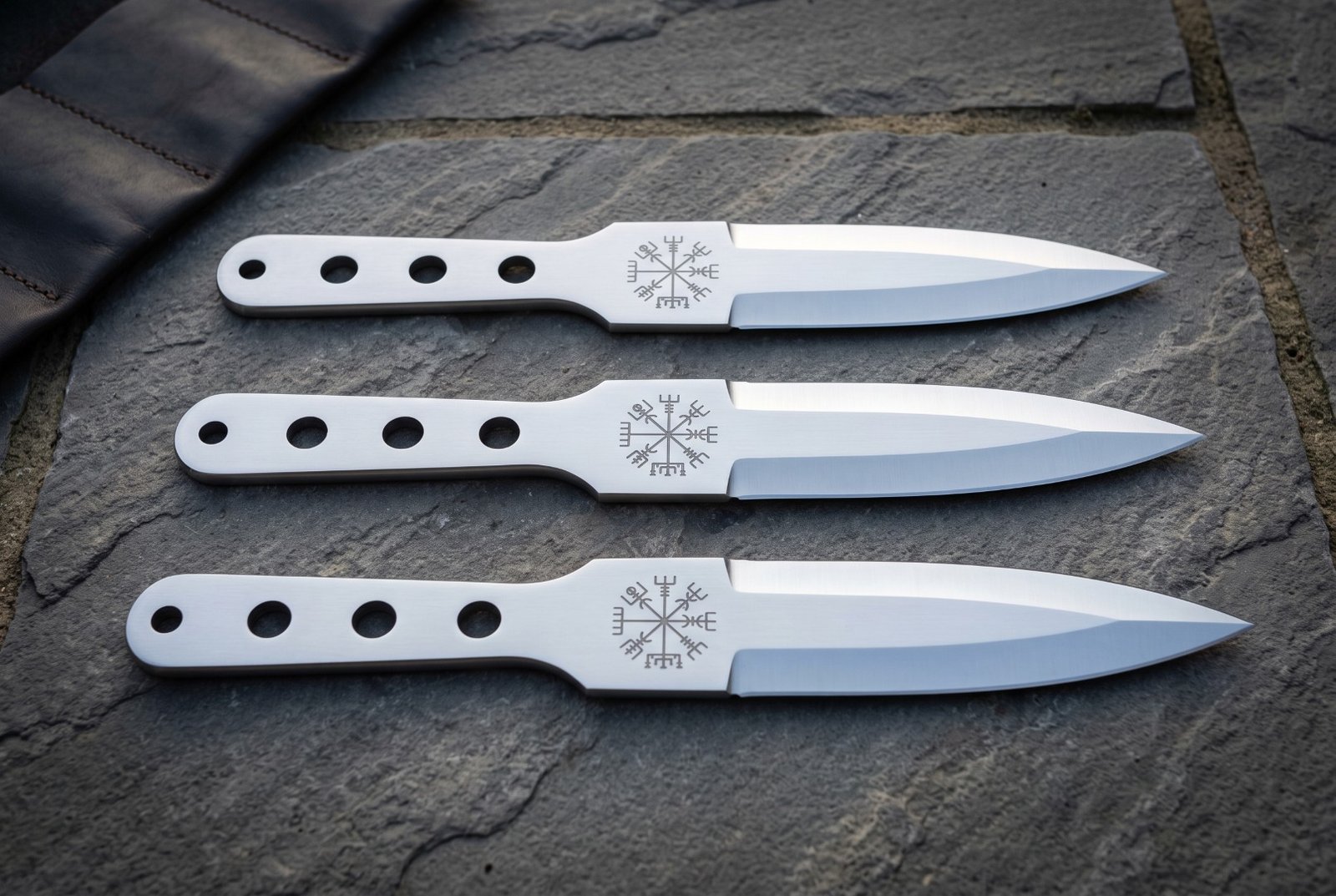 Gemini_Generated_Image_9w1d0m9w1d0m9w1d Set of three hand forged 1095 steel Viking throwing knives with skeletonised handles and Vegvisir etched blades laid on dark slate beside a leather roll