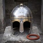 Viking spectacle helmet in polished steel with brass eye guards, engraved brow band and brass finial standing on a stone block beside a leather liner ring