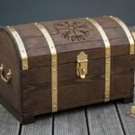 Hand made Viking storage chest in dark stained wood with brass straps, studs and end handles and a Helm of Awe engraved on the domed lid, standing on decking.
