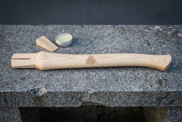 Hickory viking axe handle with a cut Valknut symbol lying on a granite bench beside its wooden wedge and a small tin of finishing wax