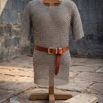 Butted mild steel chainmail hauberk with short sleeves and a thigh length skirt, hung on a wooden stand with a leather belt and a folded gambeson.