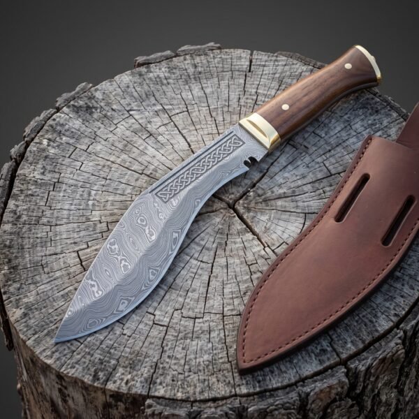 Hand forged Damascus kukri with a flowing pattern welded blade, etched knotwork panel, brass bolster and rosewood handle, on a weathered tree stump beside a leather sheath.