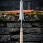 Hand forged Damascus winged spear with swept lugs, brass ferrule and pale ash haft standing against a mossy dry stone wall beside a tan leather head sheath