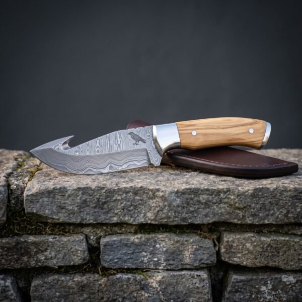 Hand forged Damascus gut hook knife with olive wood handle, engraved raven and brown leather sheath resting on a mossy limestone wall