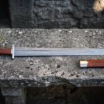 Hand forged damascus viking sword with a wide fullered blade, five lobed steel pommel and brown leather wrapped grip lying on a lichen covered stone bench beside its leather scabbard
