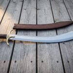 Hand forged scimitar sword with a bright polished deep curved 1095 blade, brass cross guard and dark rosewood grip lying on weathered decking boards beside its brown leather scabbard