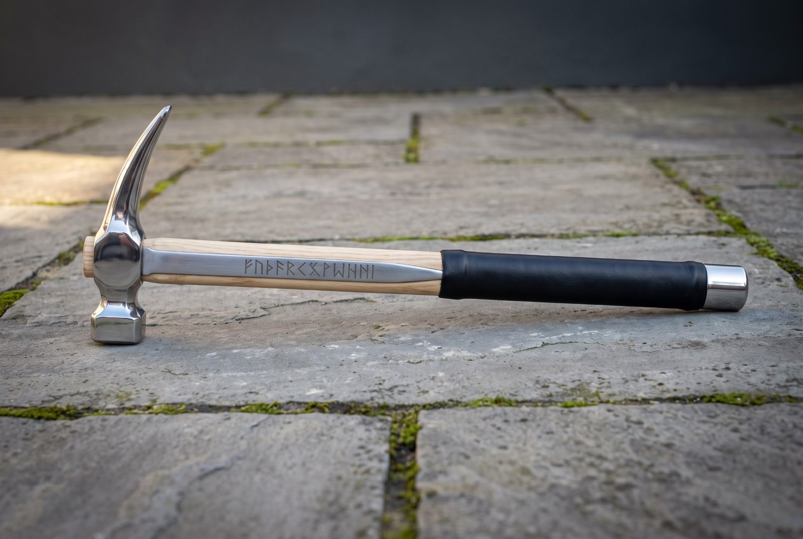 Gemini_Generated_Image_clcy3kclcy3kclcy Hand forged steel rip claw hammer with a polished head, hickory handle carrying a cut rune line, black rubber over grip and a steel end cap on stone paving.