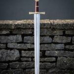 Hand forged arming sword with a brass wheel pommel and oxblood leather grip standing point down against a dry stone wall beside its red and black scabbard