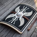 Curved roman shield painted with white wings and thunderbolts on black, domed steel boss and leather grip, laid on grey decking with its fitting kit