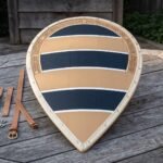 Hand made wooden kite shield painted with navy and cream bands, tooled knotwork border and rawhide rim, laid on a garden table beside its leather strap set