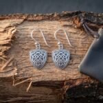 Pair of hand made Celtic earrings with shield shaped knotwork drops and beaded borders on silver hook wires lying on split birch beside a grey leather pouch