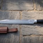 Hand forged Damascus seax knife with a wolf head etch and dark buffalo horn handle lying on stone flagstones beside a brown leather belt sheath in warm light