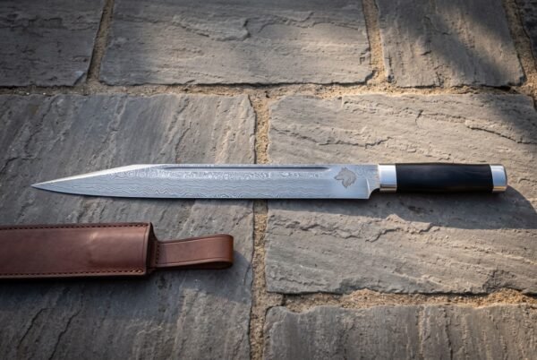 Hand forged Damascus seax knife with a wolf head etch and dark buffalo horn handle lying on stone flagstones beside a brown leather belt sheath in warm light