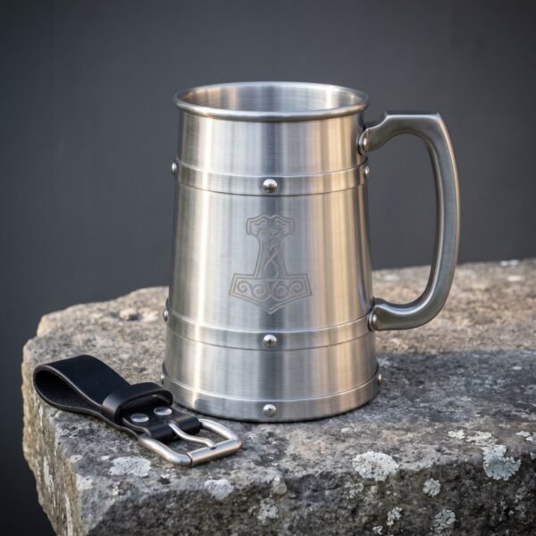 Brushed stainless steel viking tankard etched with a Mjolnir hammer and set with riveted bands, standing on a stone slab beside its black leather belt hanger