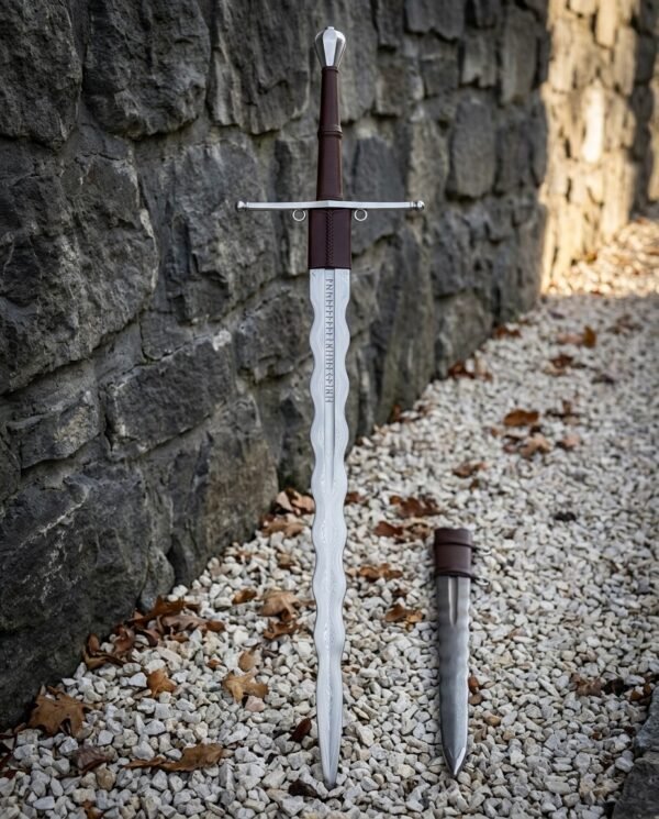Hand forged viking longsword with a flame bladed edge and rune etched ricasso standing against a stone wall on white gravel beside its matching flame bladed dagger