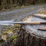 Hand forged 1095 boar spear with hooked lugs and a mirror polished head lying on a cut tree stump beside a brown leather sheath on a woodland track