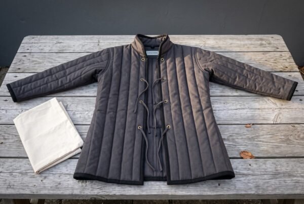 Hand made padded gambeson in charcoal cotton canvas with vertical channel quilting, standing collar and brass grommet cord lacing laid flat on a weathered grey timber table