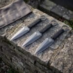 Hand forged Viking throwing knife set of three satin blades with an etched Valknut and black paracord grips laid on a sunlit stone wall beside a leather roll