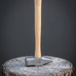 Hand forged 1095 steel cross pein forging hammer with an engraved Mjolnir on the cheek and a straight ash handle standing on an oak stump