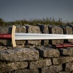 Hand forged viking sword with polished fullered blade, engraved brass knotwork guard, red leather grip and brass ring pommel lying on a dry stone wall