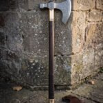 Hand forged Viking bearded axe with etched knotwork head, engraved brass collar and leather wrapped haft leaning against a lichen covered sandstone wall
