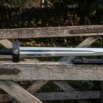 Hand forged viking sword with a Valknut engraved blackened guard and black leather wrapped grip resting on a weathered wooden farm gate beside its dark scabbard