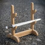 Hand made oak sword display stand with three tiers of cradles and a knotwork engraved crossbar, holding a single polished sword, standing on a gravel path.