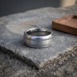 Wide silver tone Norse rune ring with an Elder Futhark centre band flanked by twisted rope borders standing on a grey stone slab beside a shallow wooden tray